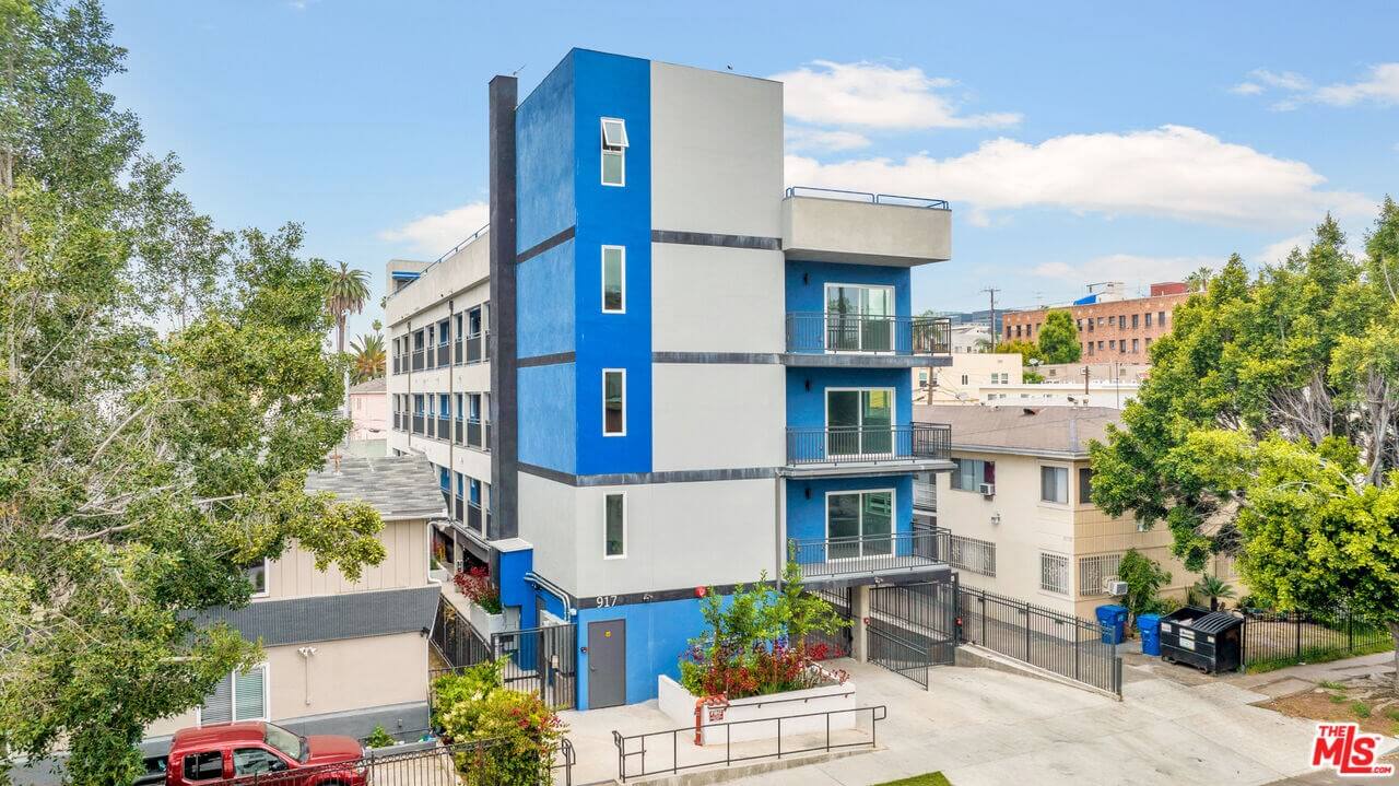 Modern blue and white apartment building surrounded by trees and neighboring houses.